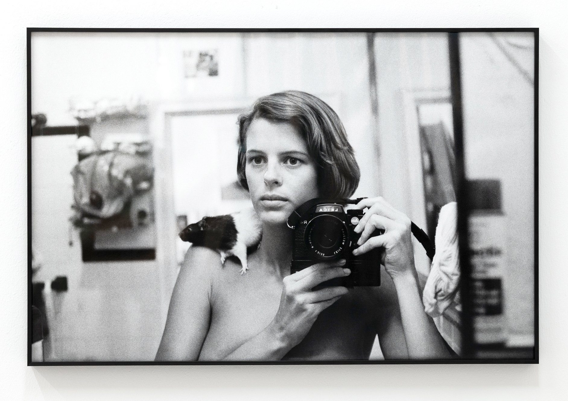 Black and white photograph of a woman taking a selfie with a camera, holding it with one hand, and a small furry dog resting on her shoulder. The woman has short wavy hair and is looking directly into the camera mirror.