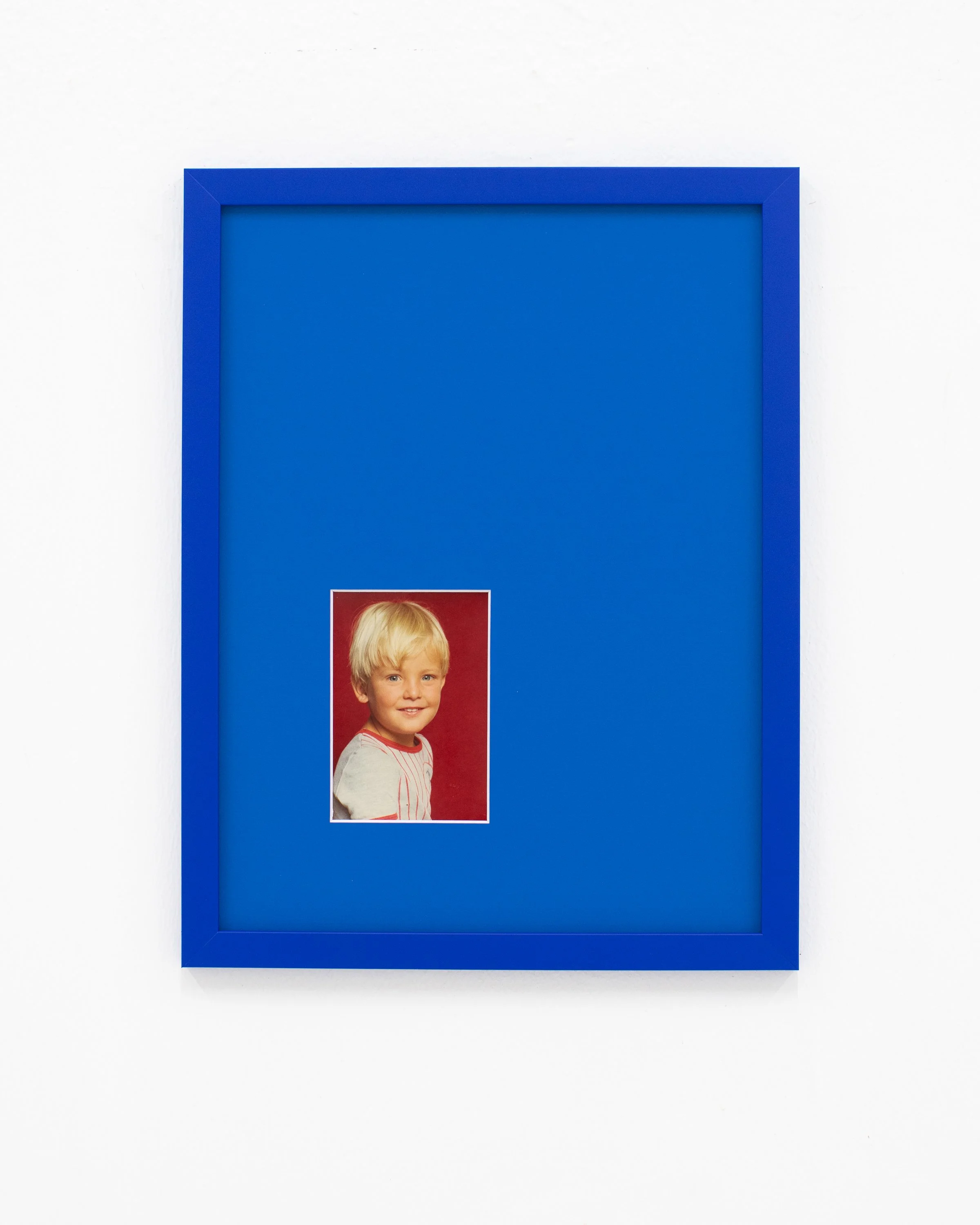 Photograph of a framed portrait of a young blonde-haired boy against a red background, mounted on a white wall and encased in a blue frame.