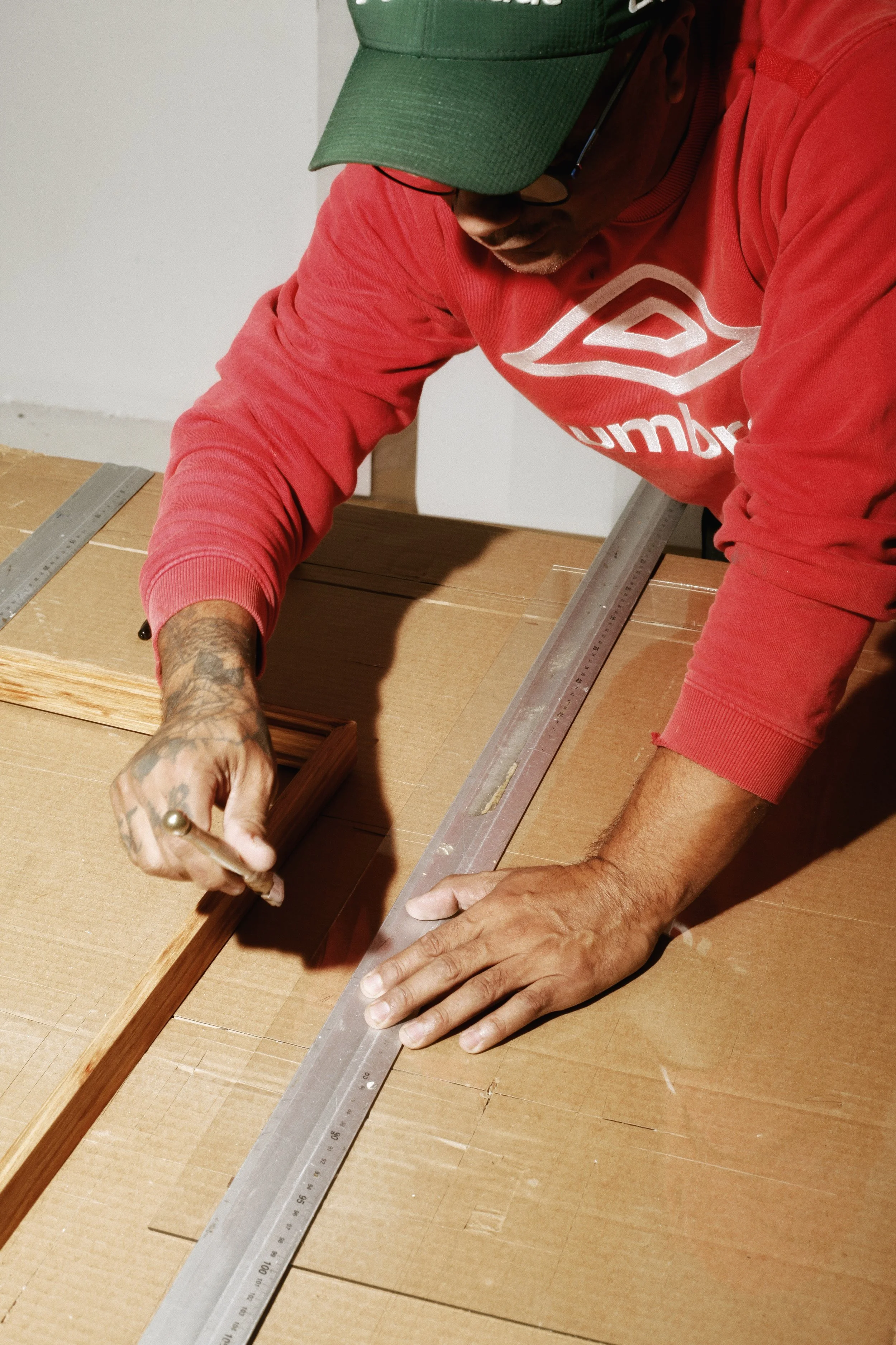 A man wearing a green cap and red sweatshirt is measuring and marking wood with a metal ruler and a pencil on a work table.