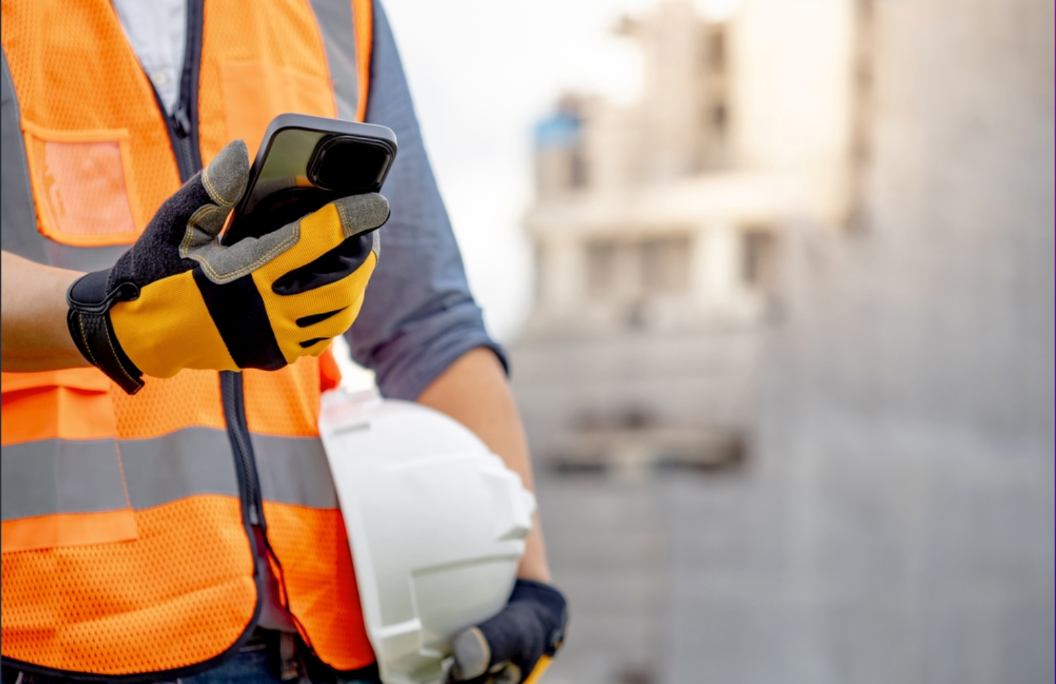 Close-up of a construction worker wearing yellow gloves, an orange safety vest, holding a smartphone and a white helmet at a construction site.
