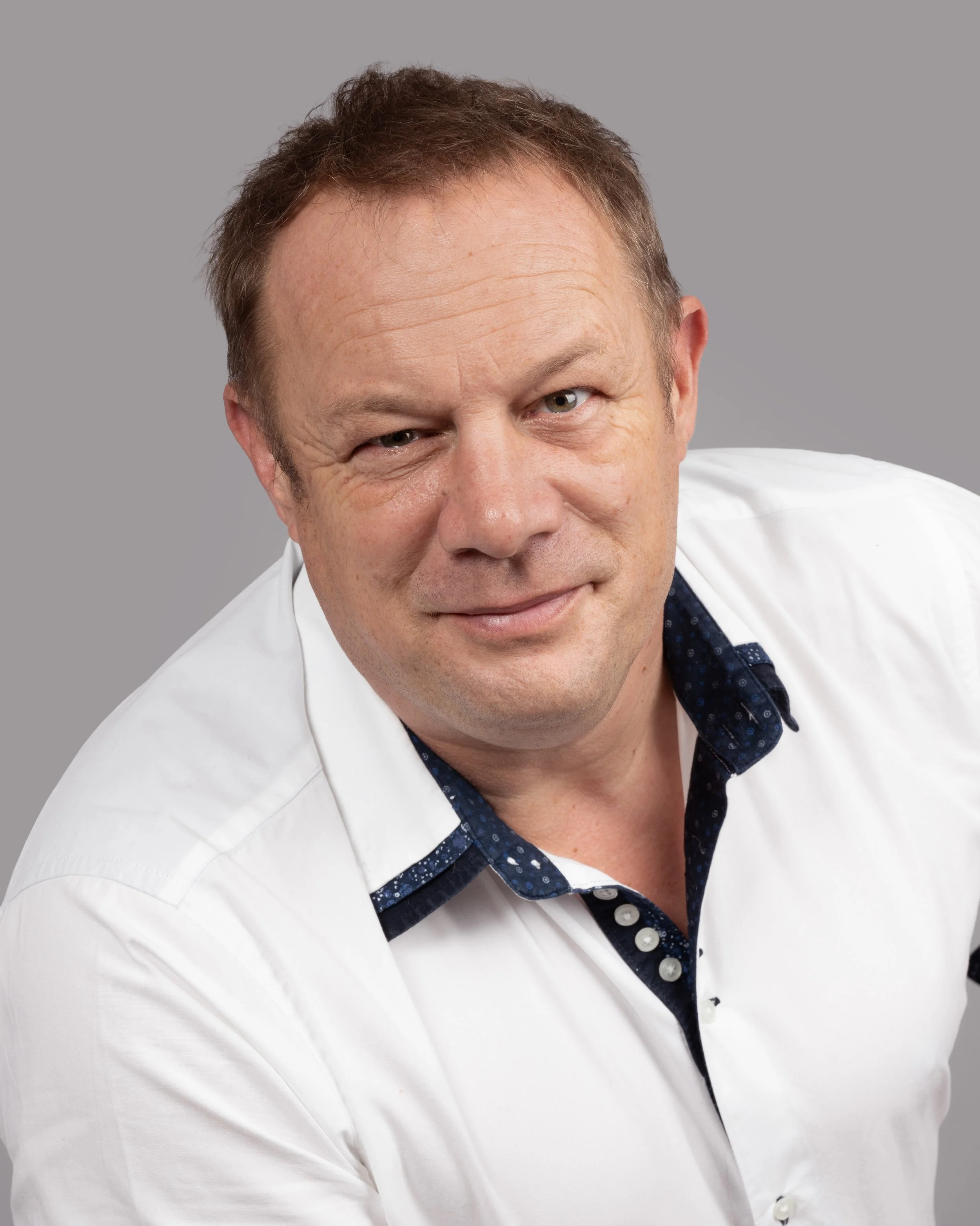 A middle-aged man with light skin and short brown hair, wearing a white shirt with dark blue patterned collar and inner lining, smiling and tilting his head slightly against a neutral gray background.