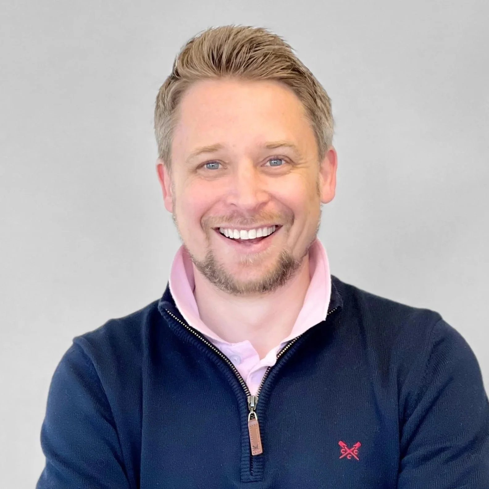 A smiling man with short blond hair, blue eyes, and a beard, wearing a light pink collared shirt and a dark zip-up jacket, standing against a plain light gray background.
