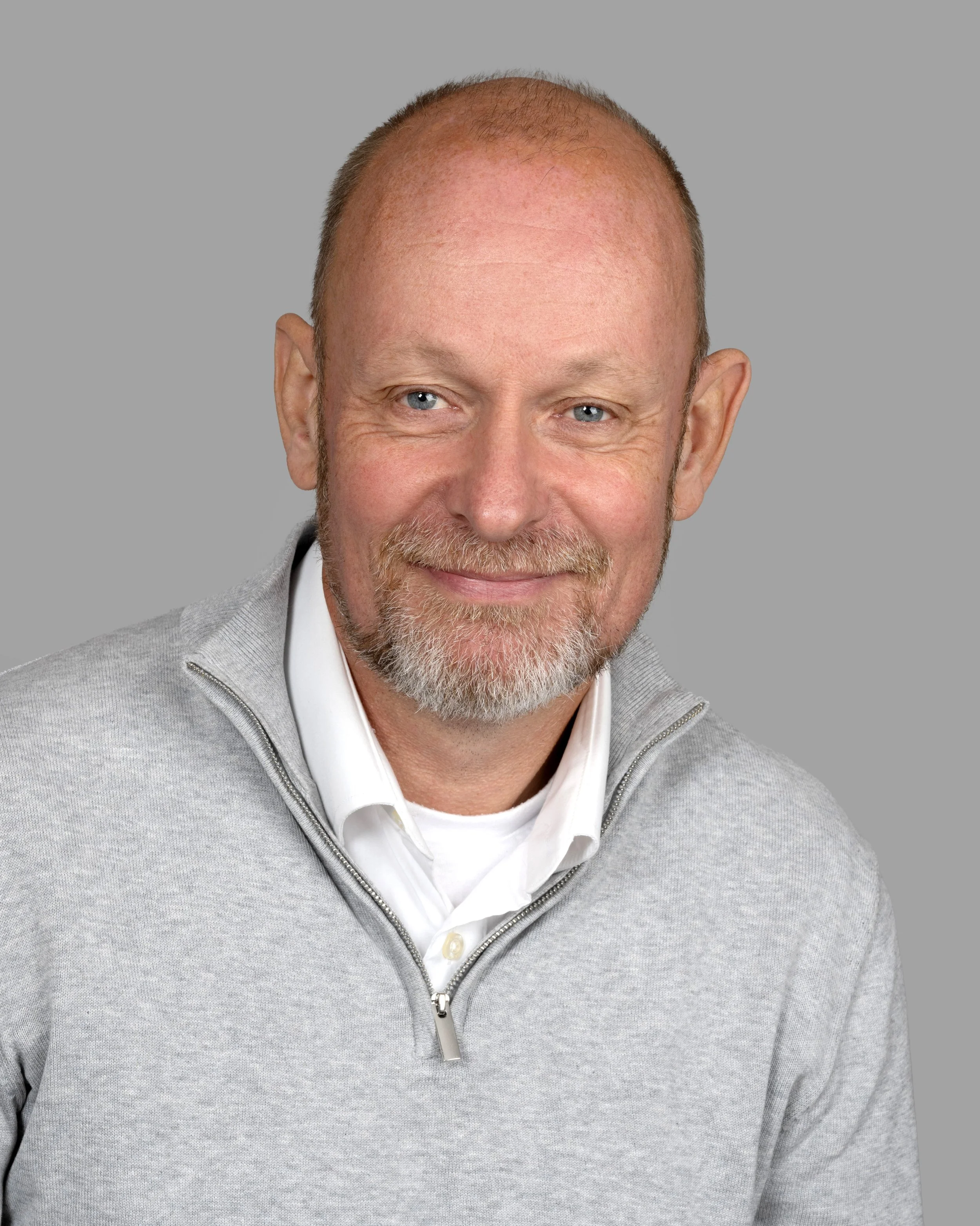 A middle-aged man with a beard and balding head, wearing a light gray zip-up sweater over a white collared shirt, smiling against a plain gray background.
