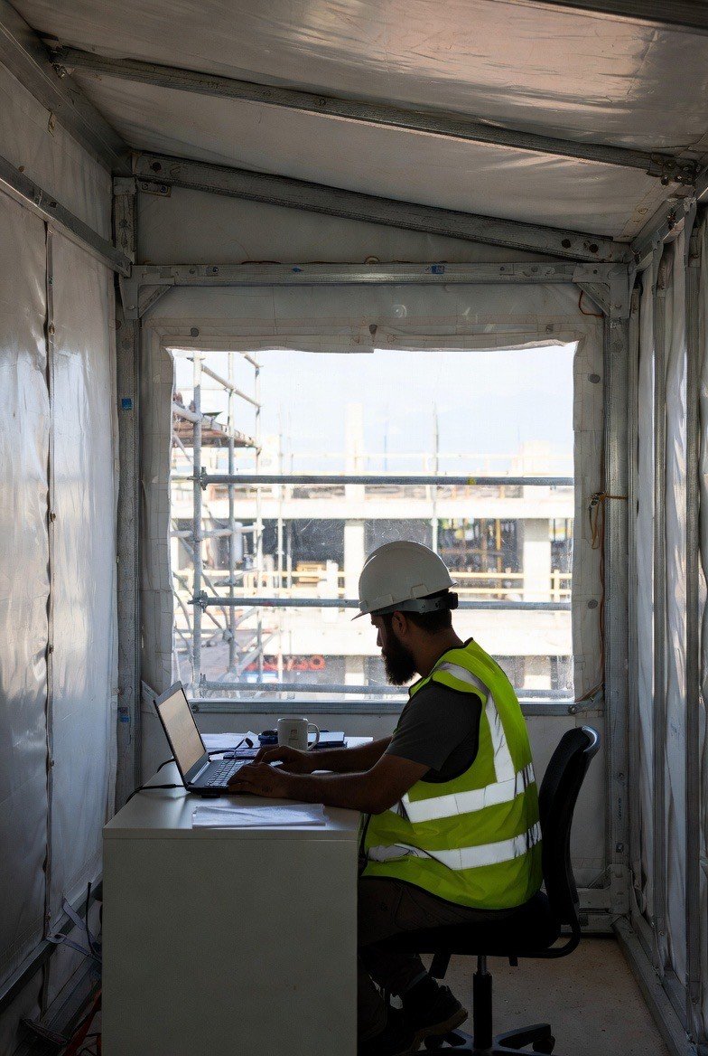 A construction worker wearing a white safety helmet and a yellow reflective vest working on a laptop inside a portable office container at a construction site.