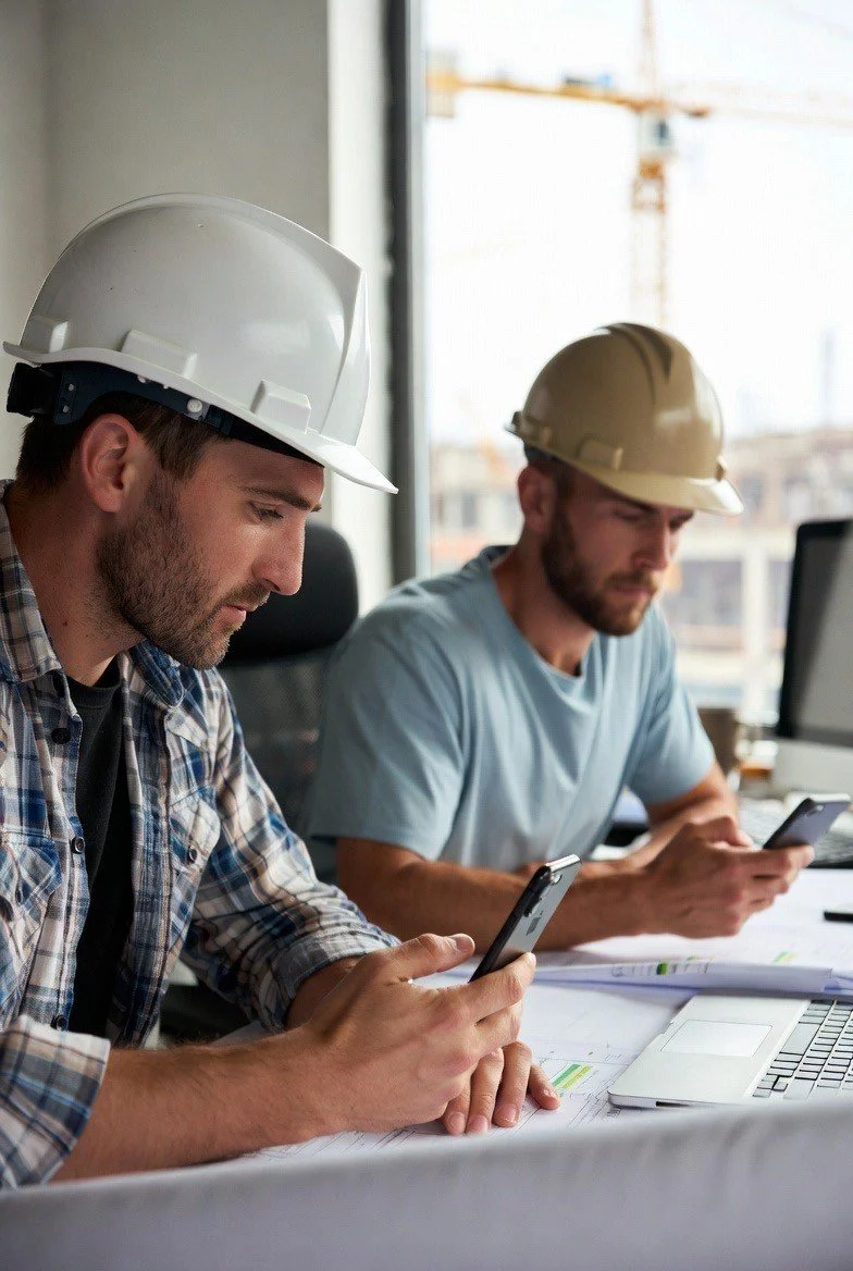 Two construction workers wearing hard hats are sitting at a desk, looking at their smartphones in an office with large windows and a city view.