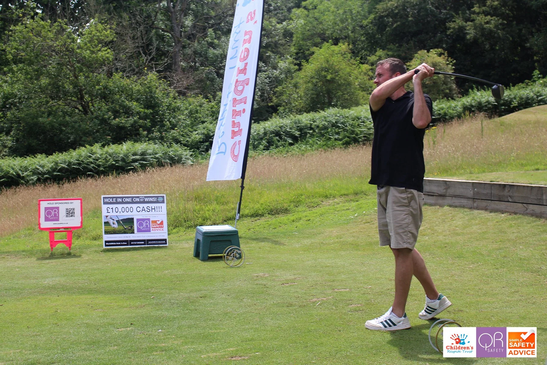 A man in black shirt and beige shorts swinging a golf club on a golf course, with a sign offering a chance to win 10,000 pounds cash in the background, along with various safety and community logos.