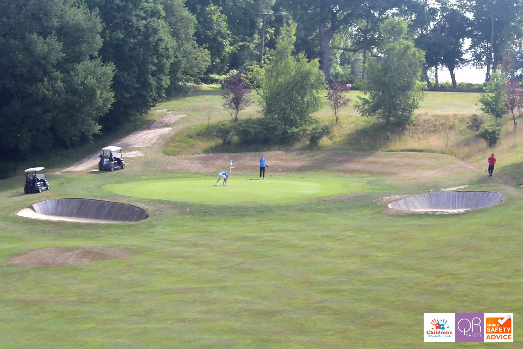A golf course with two bunkers and three golfers near the green, surrounded by trees and a dirt path.