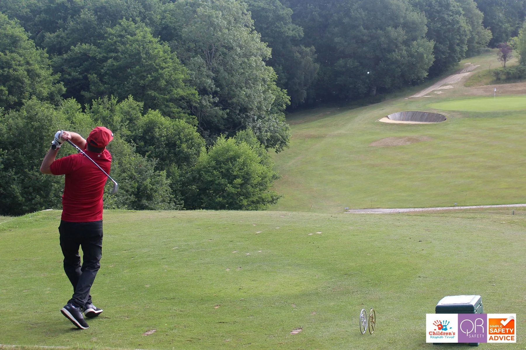 A person in a red shirt and black pants swinging a golf club on a golf course surrounded by trees, with a bunker and bushes in the distance, and two small wheels and a cooler on the ground.