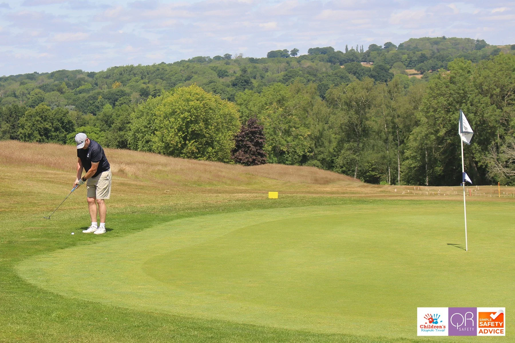 A man in a black shirt, khaki shorts, and a white cap is putting on a golf course near the green, with trees and hills in the background.