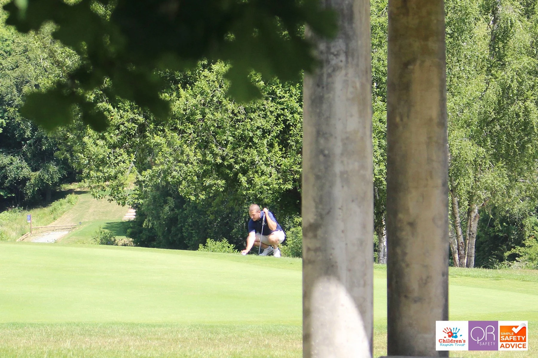 A man in a blue shirt and shorts kneeling on a golf course, preparing to putt a golf ball, with trees and a grassy landscape in the background.