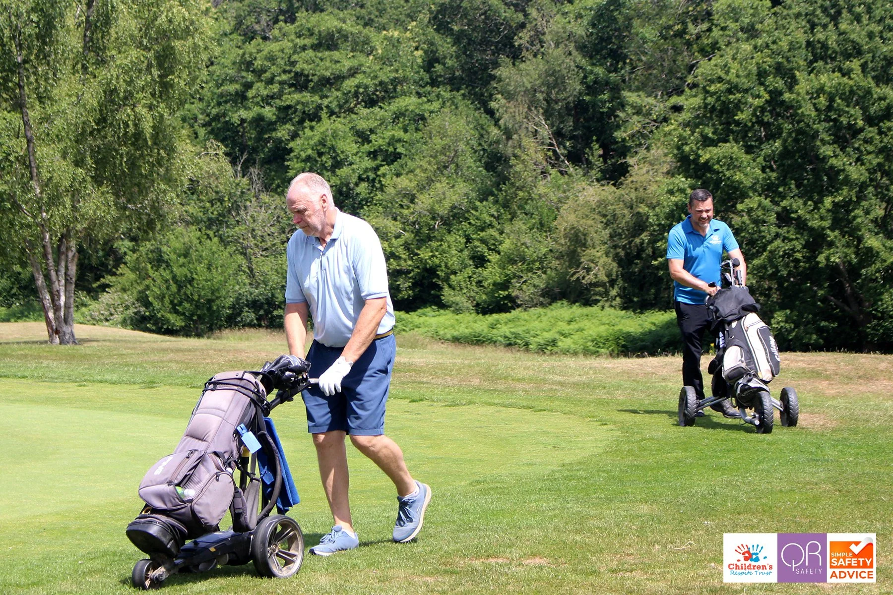 Two men are on a golf course, each pushing a golf cart with golf bags. One is an elderly man with white hair, wearing a light blue polo shirt and blue shorts. The other is a middle-aged man with black hair, wearing a bright blue polo shirt and black 