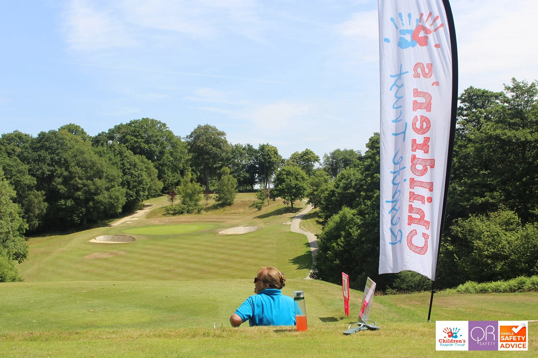 A woman sitting on a golf course near a flag with children's safety logos, trees, grass, and sand bunkers in the background.