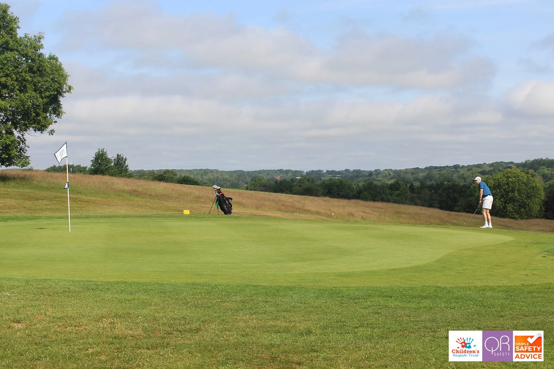 A golfer on a course preparing to putt, with a golf bag and flag in the foreground and treeline in the distance.