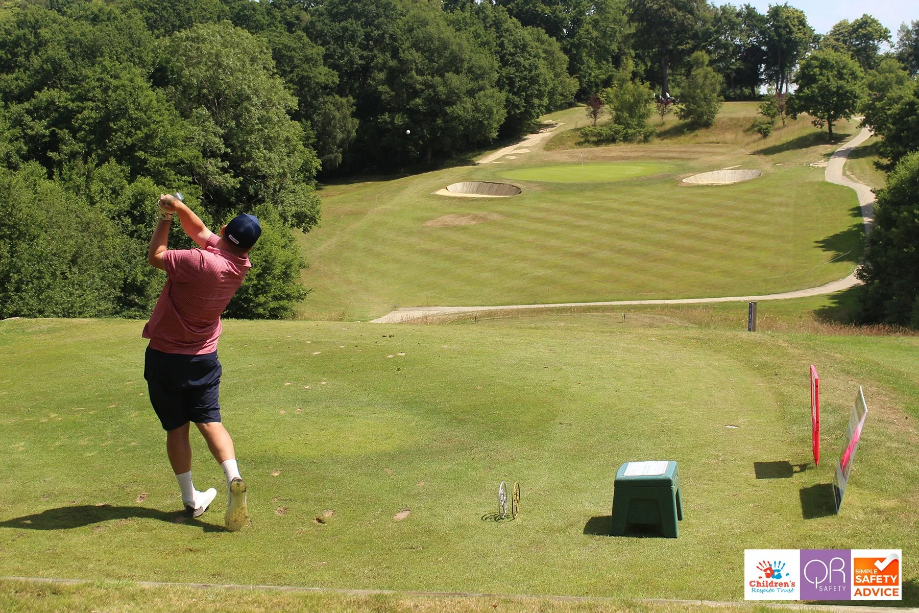 A person in a pink shirt and navy shorts swings a golf club on a golf course with trees and bunkers in the background.