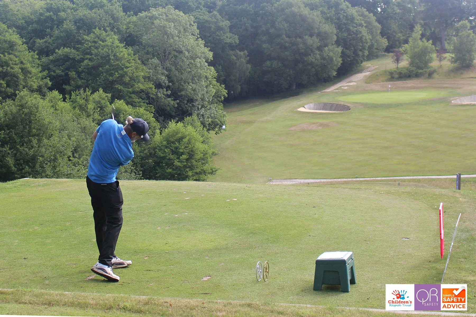 A person in a blue shirt and black pants takes a golf swing on a tee box at a golf course, with lush trees and a green in the background.