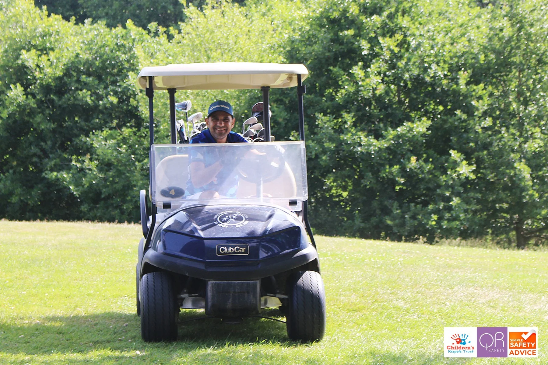 A man sitting in a golf cart on a grassy field, smiling, with a background of green trees.