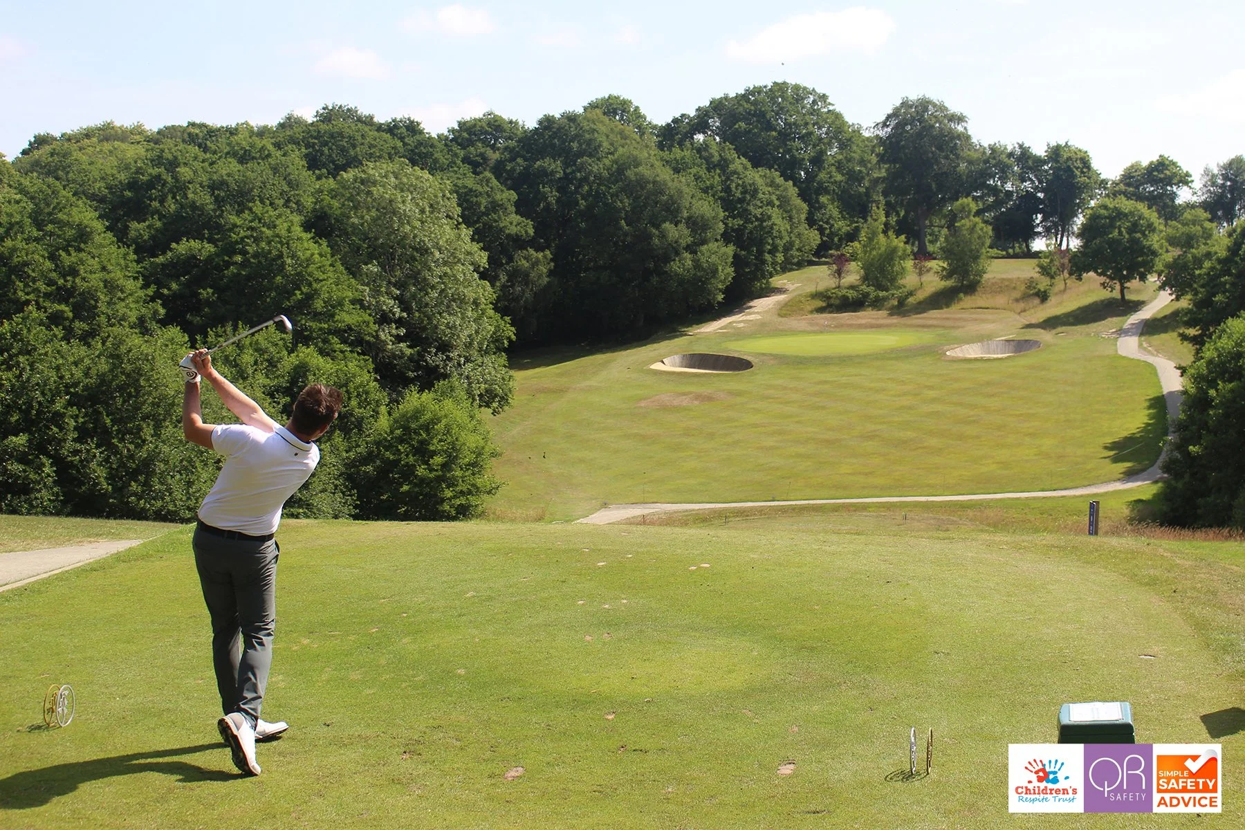 A man plays golf on a lush, green course surrounded by trees, with sand traps in the distance under a sunny sky.