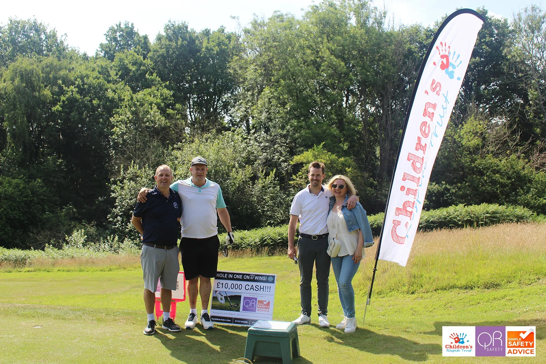 Four people standing on a golf course, smiling and posing for a photo next to a large sign and a flag, with trees in the background.
