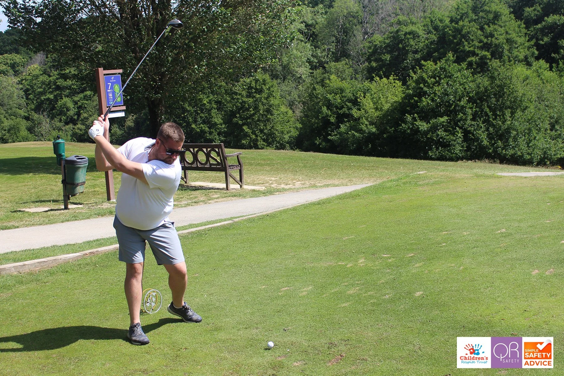 A man in a white shirt and gray shorts is swinging a golf club on a golf course. He is wearing sunglasses and is about to hit a golf ball on the green, with trees and a bench in the background. There are safety and advocacy logos in the bottom right 