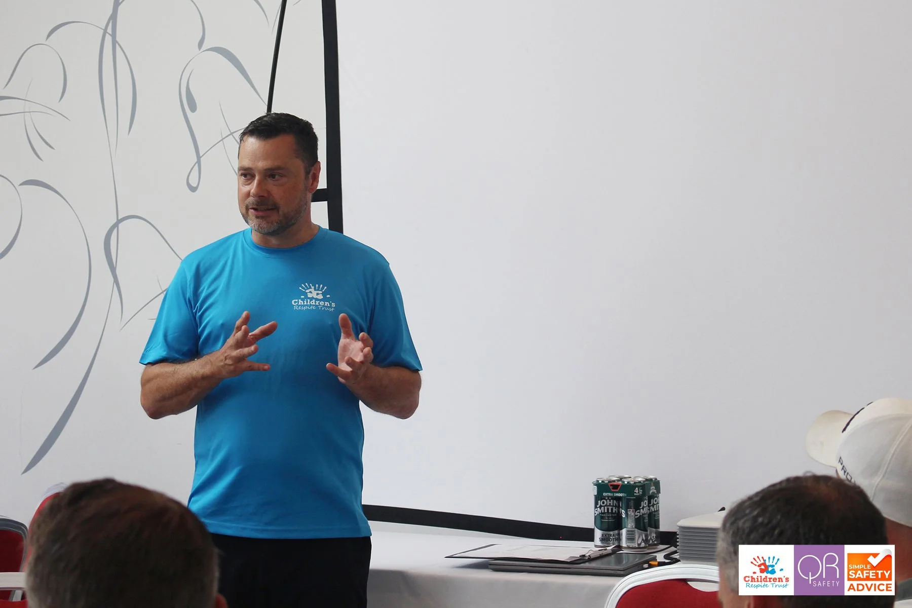 A man in a bright blue t-shirt giving a presentation in a room with white walls and a black and white mural. There are cans of John Smith soda and stacks of paper on a table nearby, with an audience in front.