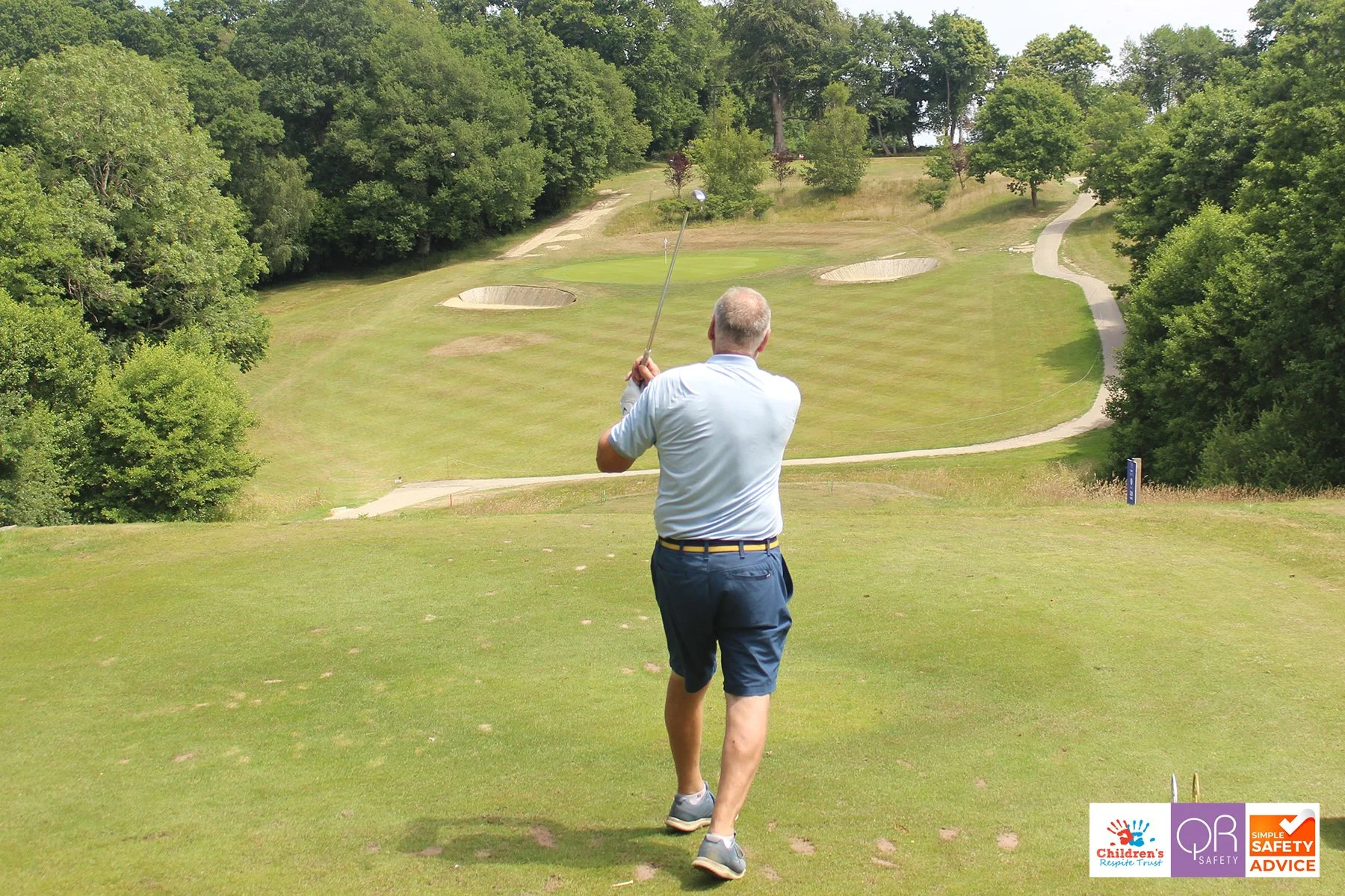 A man in a light blue shirt and navy shorts is playing golf on a lush green golf course with trees and sand traps, viewed from behind as he swings his club.