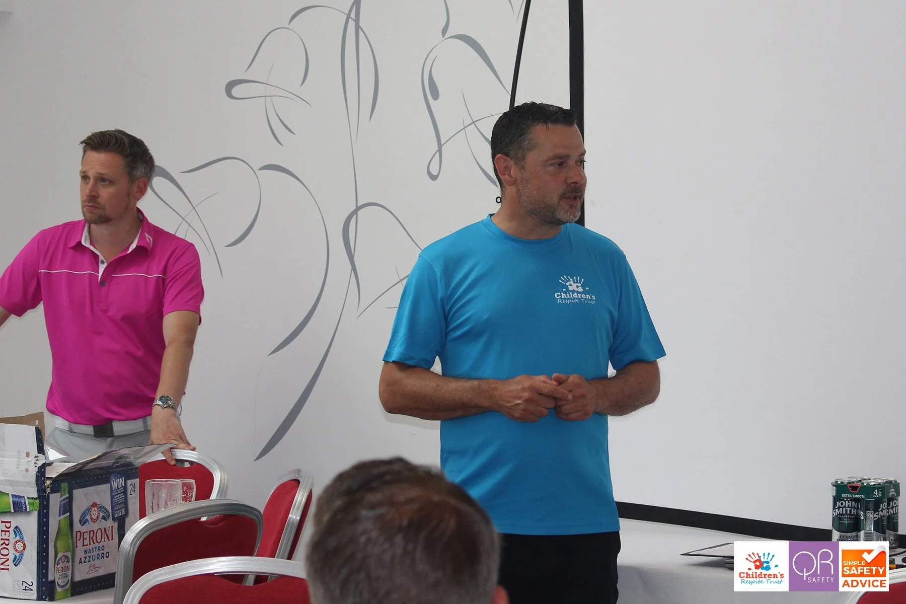 Two men standing in a room with a white wall, one wearing a pink polo shirt and the other wearing a blue T-shirt, which has a logo for Children's Response Trust. There are tables and chairs with boxes and cans of drinks on the tables. A person’s head