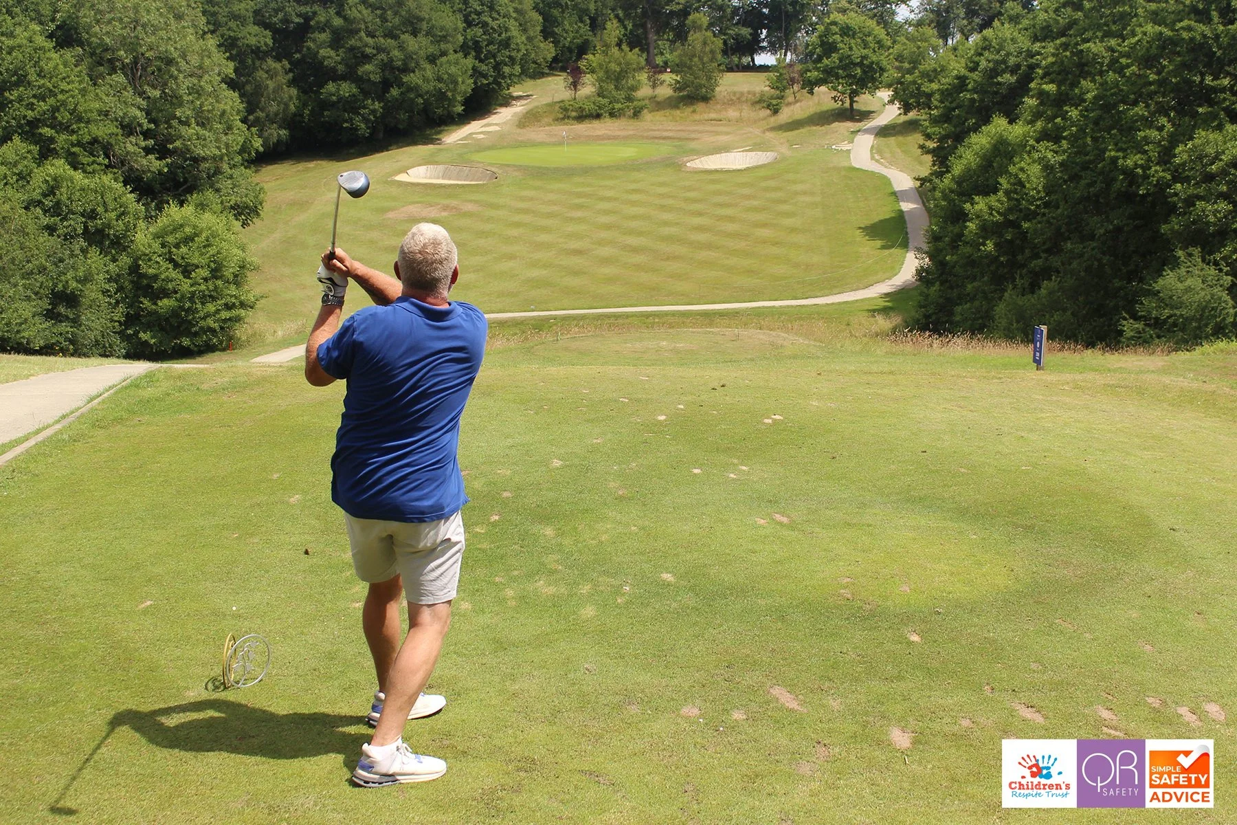 A man with gray hair in a blue shirt and beige shorts is swinging a golf club on a grassy golf course. The course has trees and a winding path, with sand bunkers near the green which has a flag, and a small sign on the right. The bottom right corner 