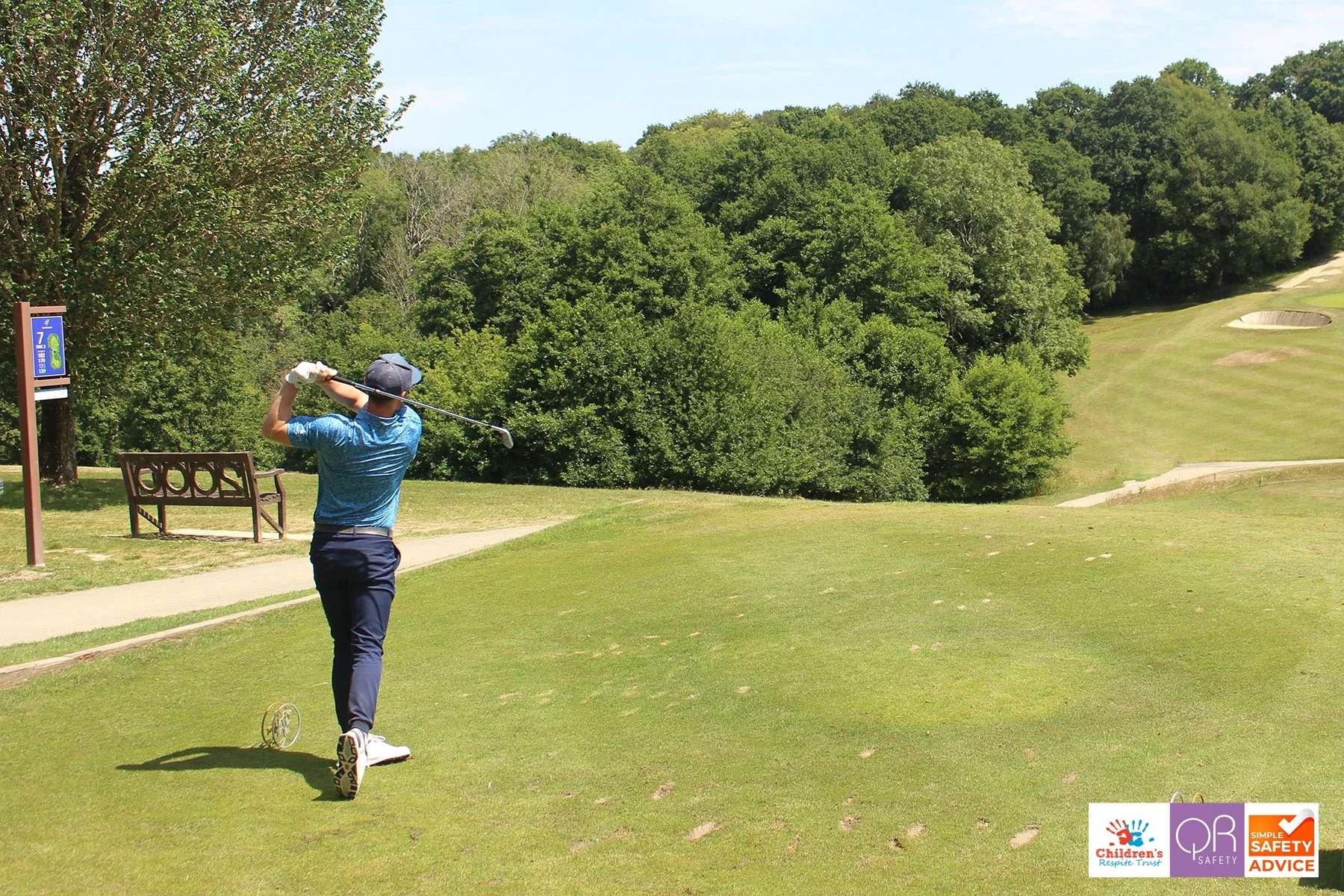 A person playing golf on a sunny day, swinging a golf club on a golf course with green grass and trees in the background.