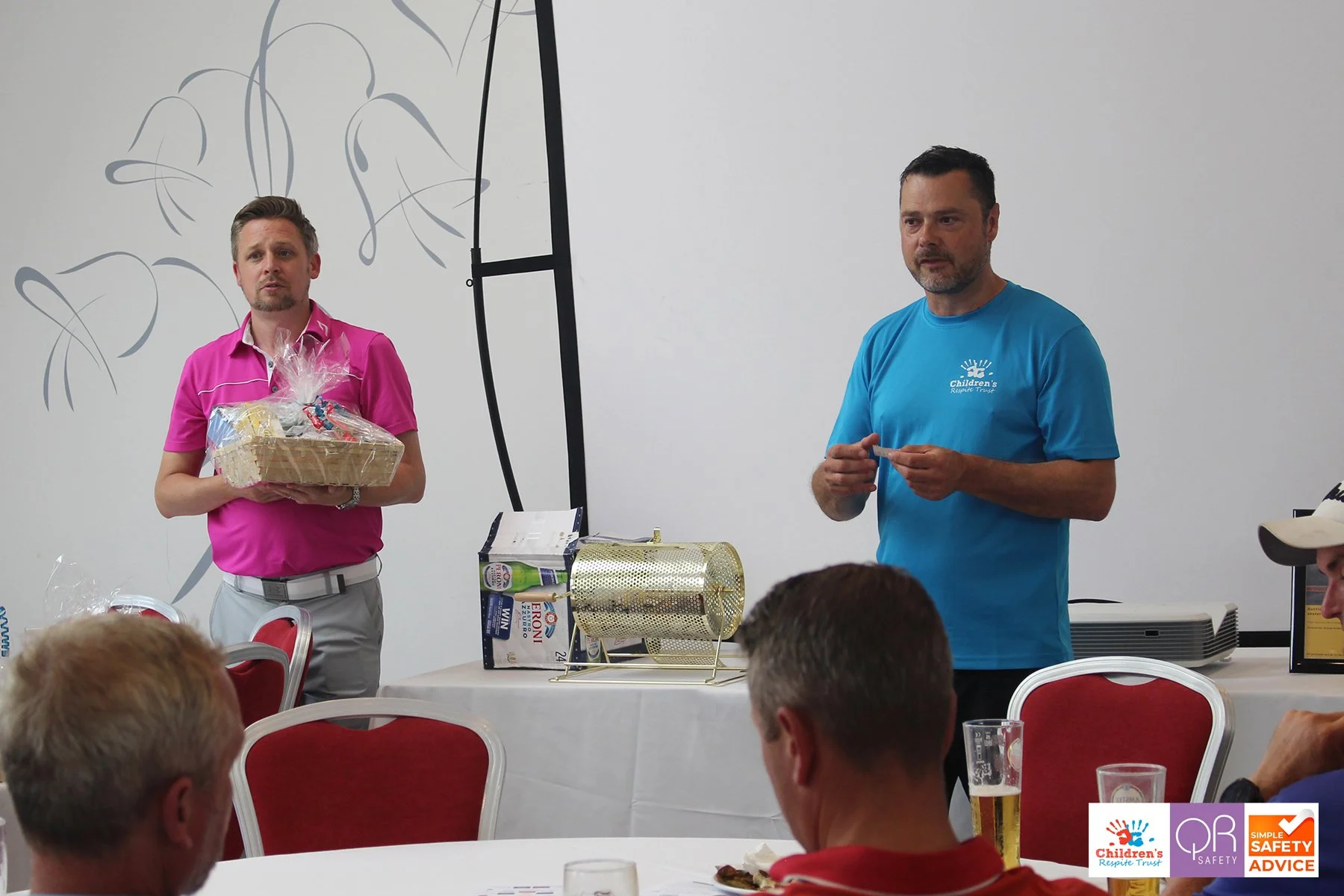 Two men standing in front of an audience at a presentation or event, with a white wall and decorative black lines in the background. The man on the left wears a pink shirt and holds a gift basket wrapped in cellophane, while the man on the right wear