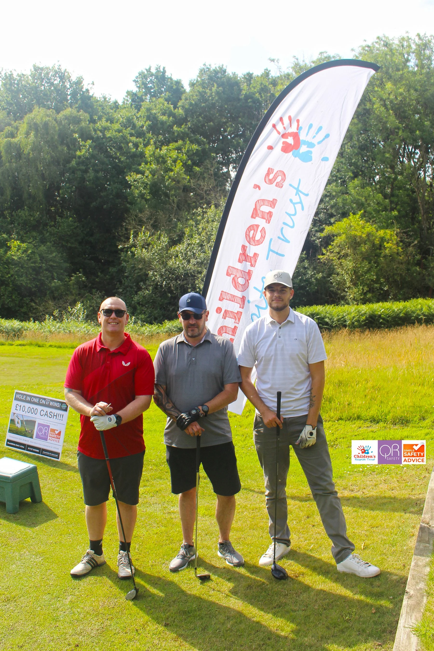 Three men standing on a golf course holding golf clubs, with a golf tournament banner and a sign promoting a cash prize in the background.