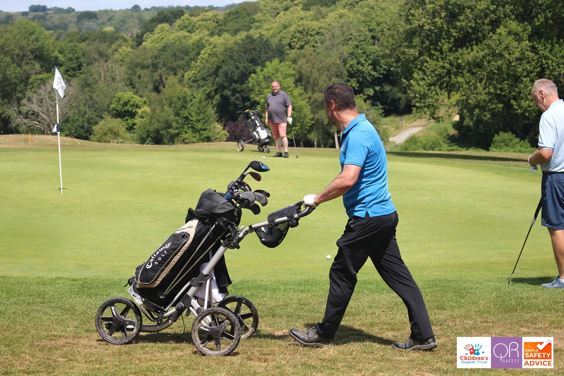 Three men on a golf course, one pushing a golf cart and the other two preparing to hit golf balls. Green trees in the background.