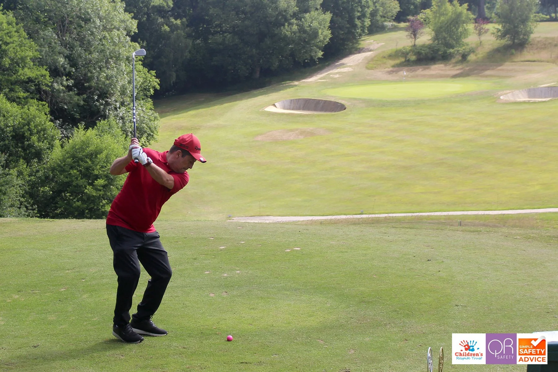 A man in a red shirt, black pants, and a red cap preparing to hit a golf ball with a golf club on a golf course.