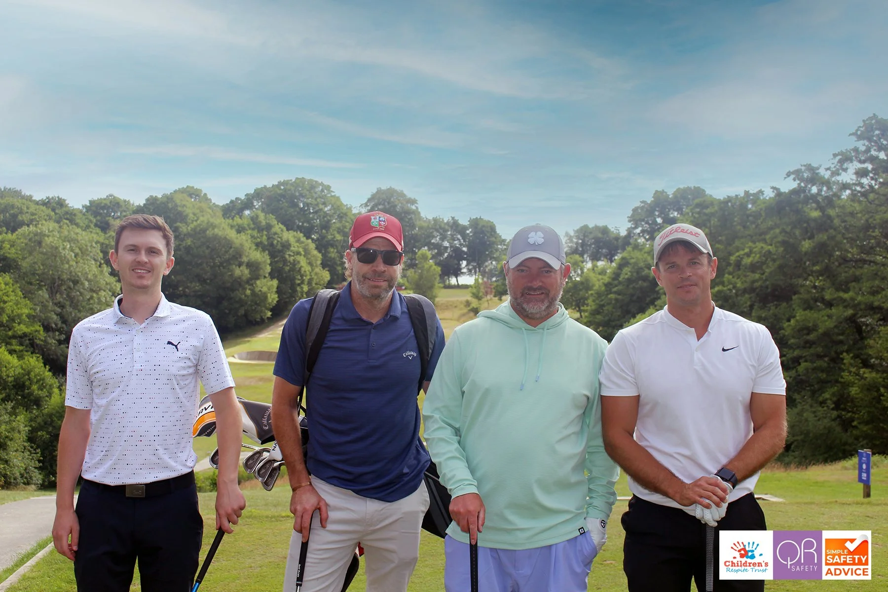 Four men standing on a golf course with trees and a partly cloudy sky behind them, holding golf clubs.