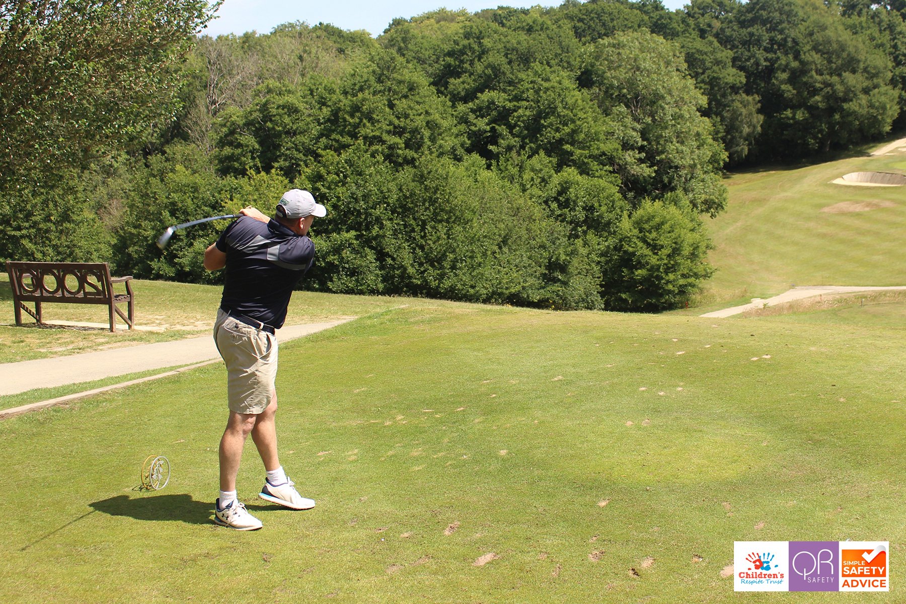 Man in black shirt and beige shorts swinging a golf club on a golf course surrounded by trees.