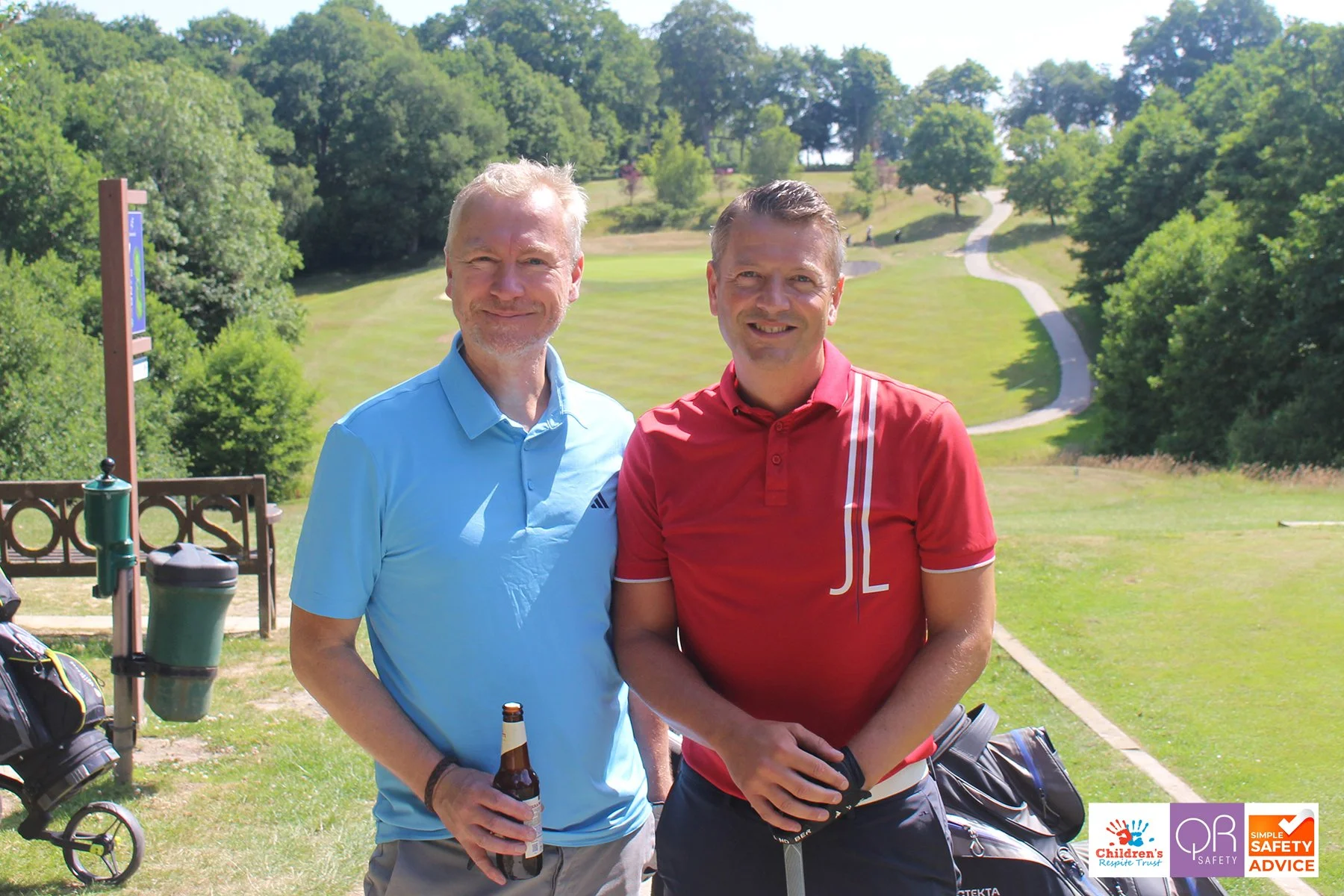 Two men standing on a golf course with trees and a pathway in the background, one holding a beer bottle and the other holding golf gloves, smiling at the camera.