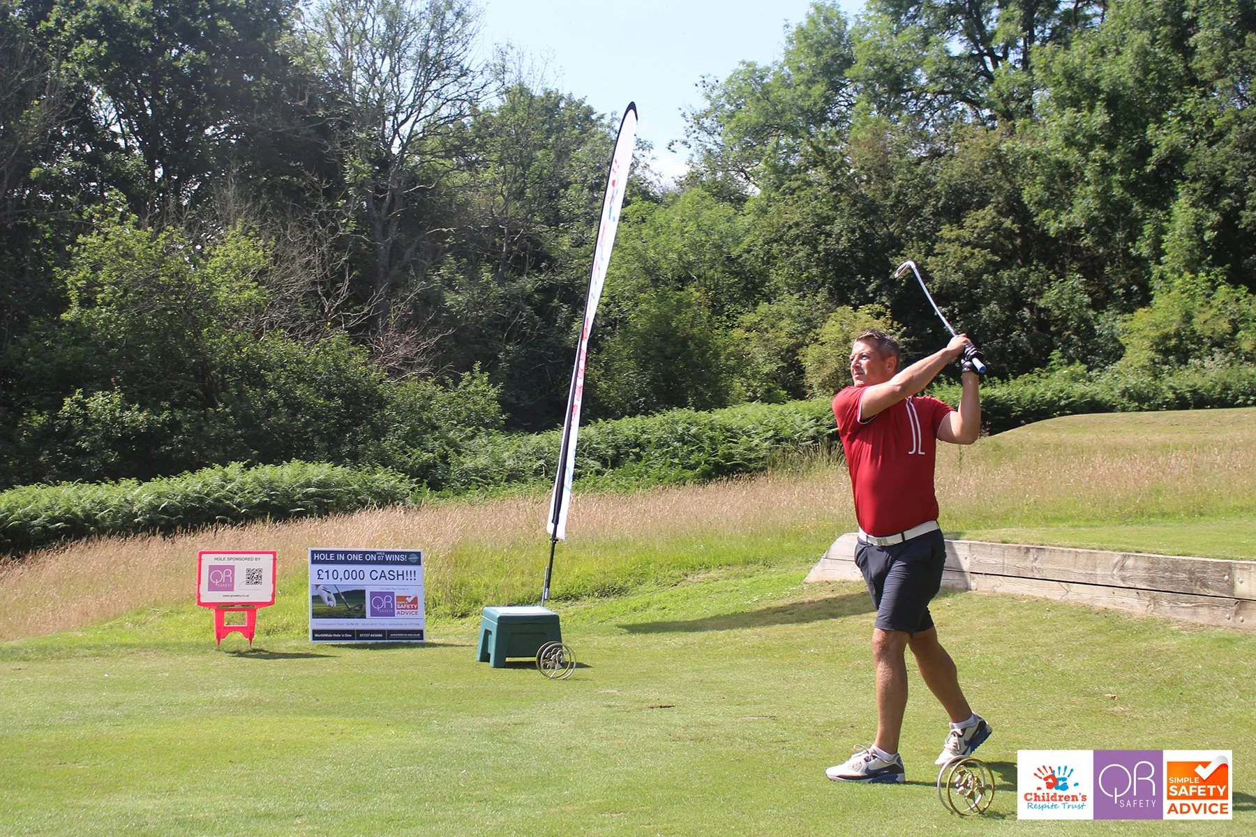A man in a red shirt and shorts playing golf on a sunny day at a golf course, with trees and advertisements visible in the background.