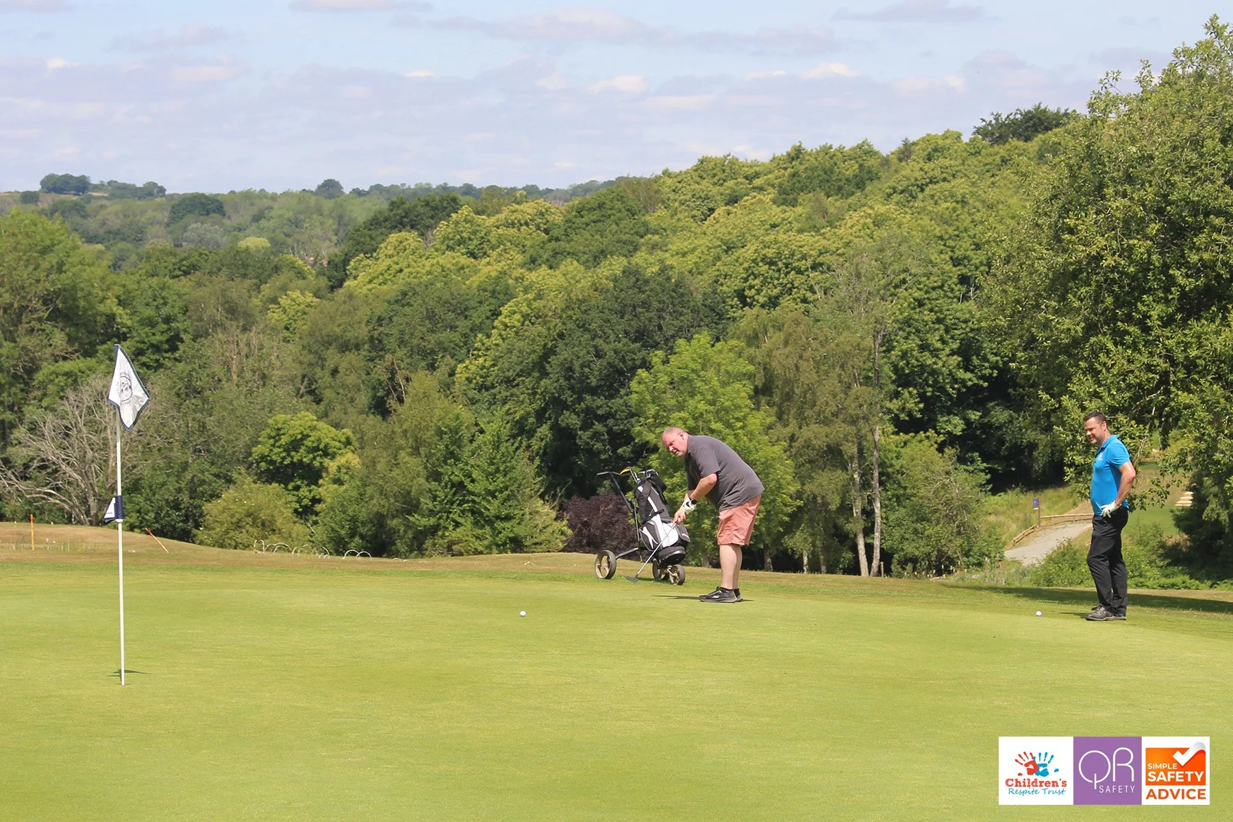 Two men on a golf course, one preparing to putt, with a flagstick on the green, surrounded by trees and hills in the background, and logos for Children's Respire Trust, OP Safety, and Simple Safety Advice in the bottom right corner.