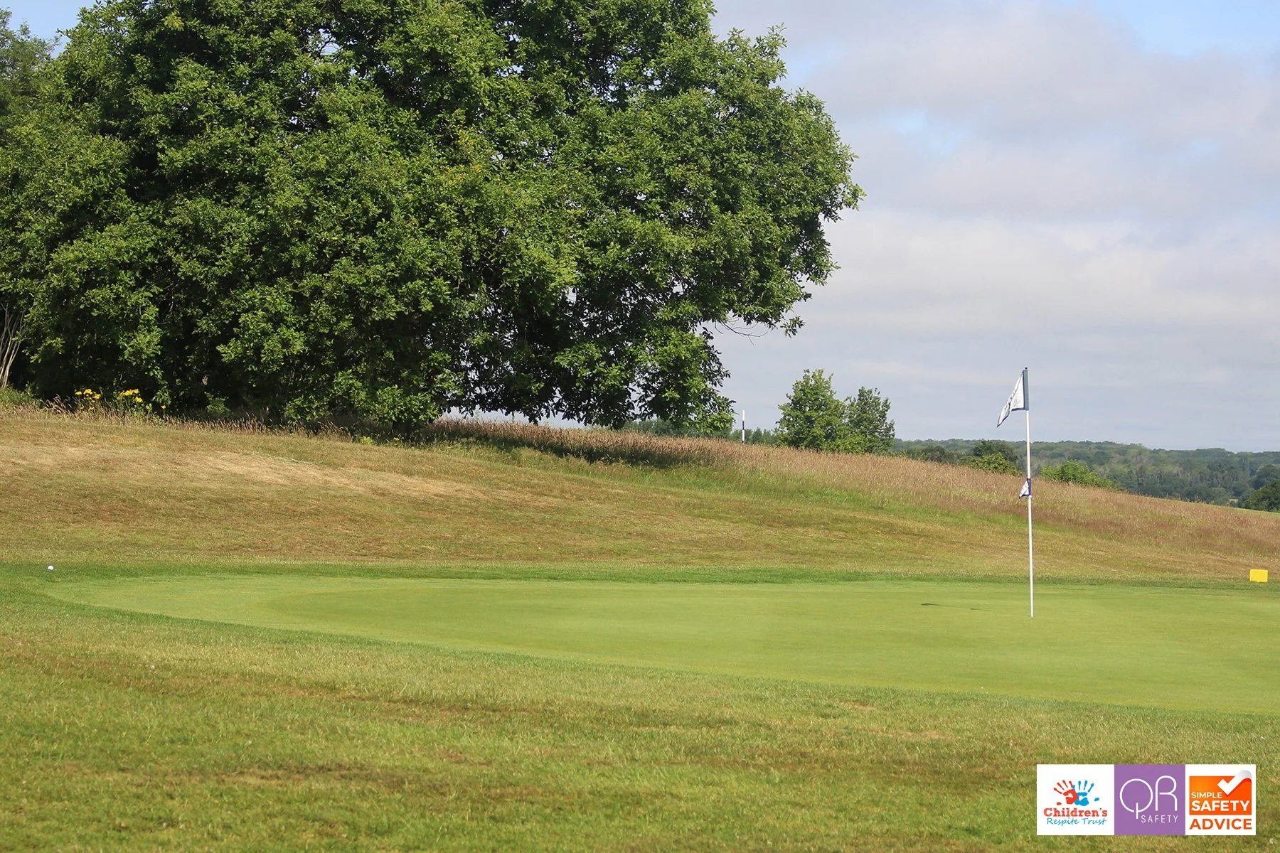 A golf course with a green, flagstick, and hole, surrounded by grass, a hillside, and trees under a partly cloudy sky.