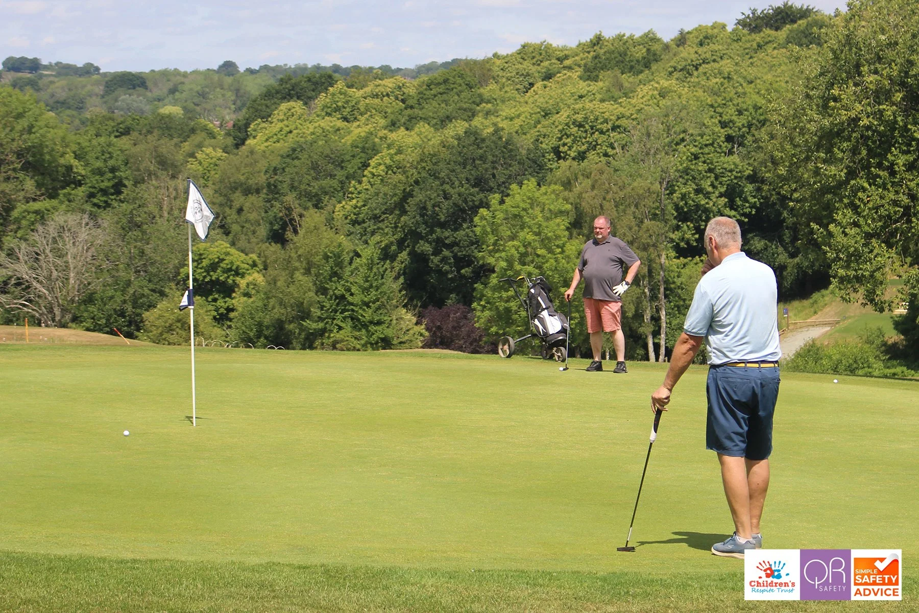 Two men playing golf on a course with a lush green landscape and trees in the background. One man is preparing to putt while the other stands nearby with a golf cart. Safety and children’s charity logos are in the bottom right corner.