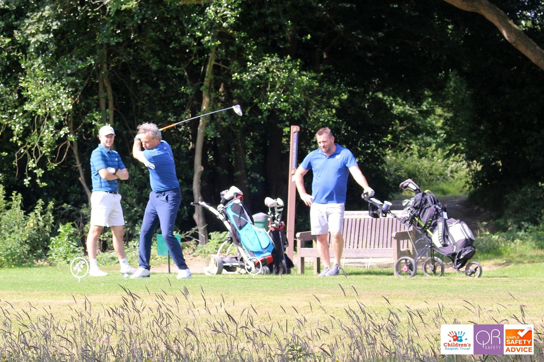 Three men playing golf on a course surrounded by trees and grass, with golf bags and equipment nearby.