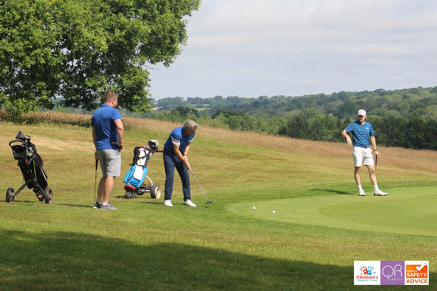 Three men playing golf on a lush golf course, with two of them preparing to hit the ball, and one watching, with golf bags and clubs nearby, under a large tree and a partly cloudy sky.