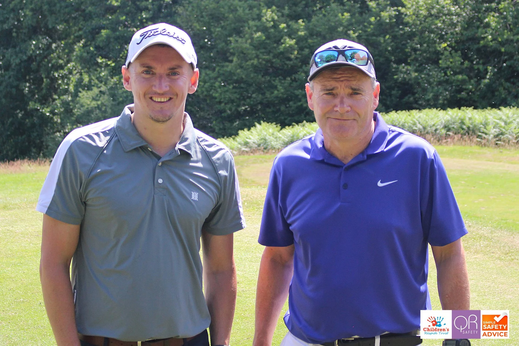 Two men standing on a golf course, smiling. The man on the left wears a gray golf shirt and a white cap, while the man on the right wears a blue polo shirt and sunglasses on his head.