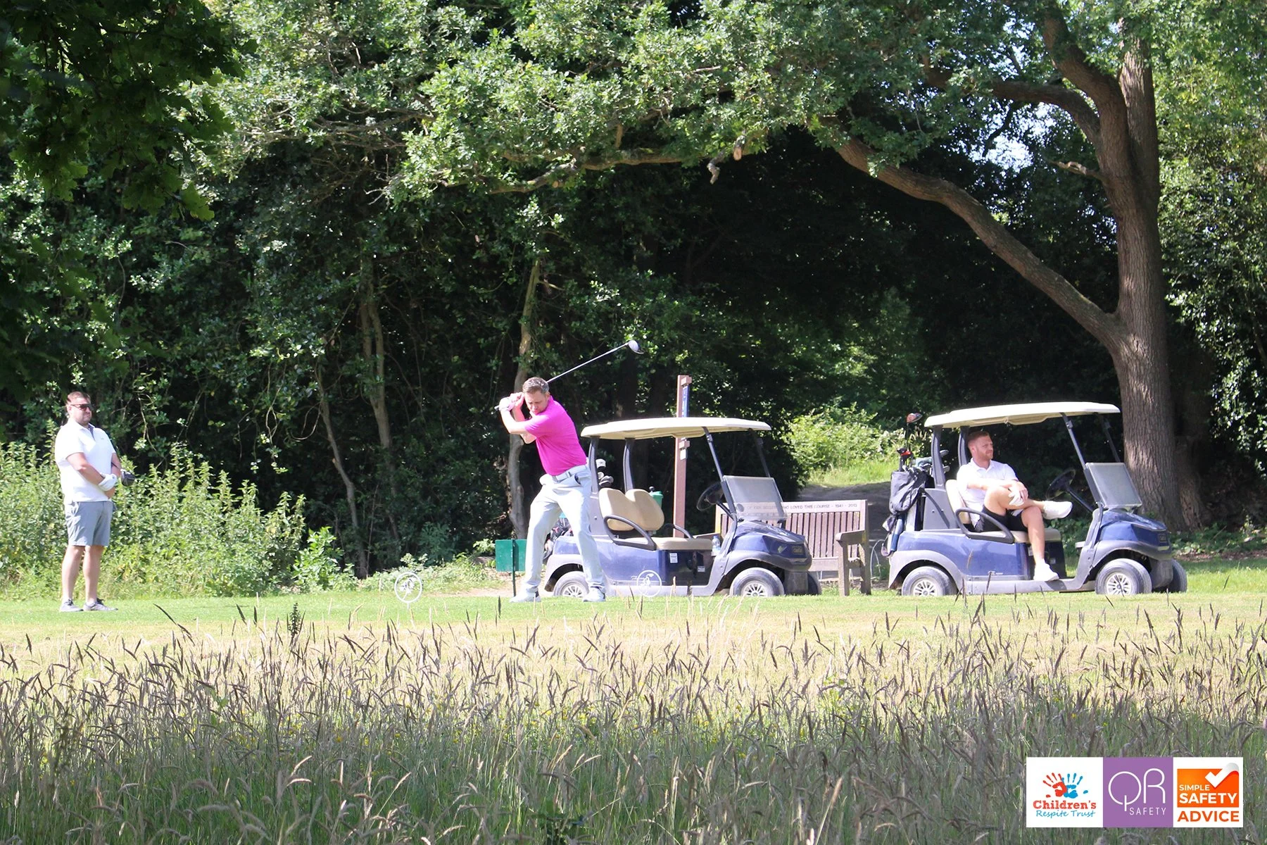 People playing golf on a course with two golf carts parked nearby, surrounded by large trees and greenery.