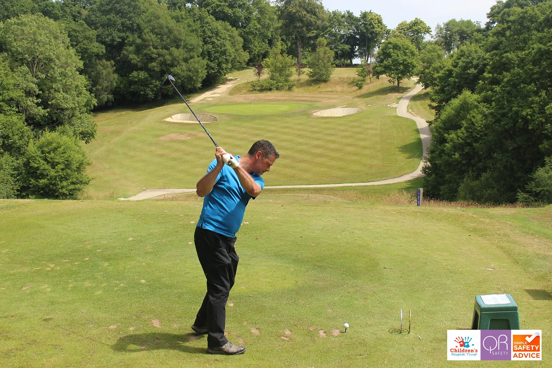 A man in a blue shirt and black pants swinging a golf club on a golf course. The course is lush green with trees and sand traps in the background. There are safety logos in the bottom right corner.