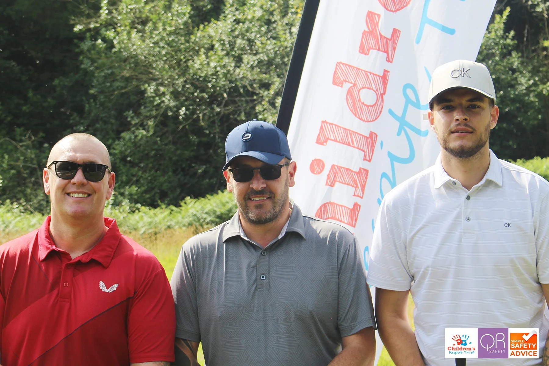 Three men standing outdoors in front of a white banner with red and blue text, all wearing golf shirts and sunglasses, with trees in the background.