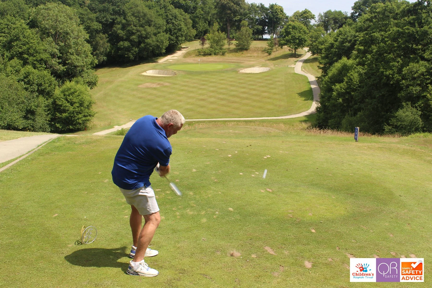 A man in a blue shirt and shorts hitting a golf ball on a teeing ground at a golf course with trees and a putting green in the background.