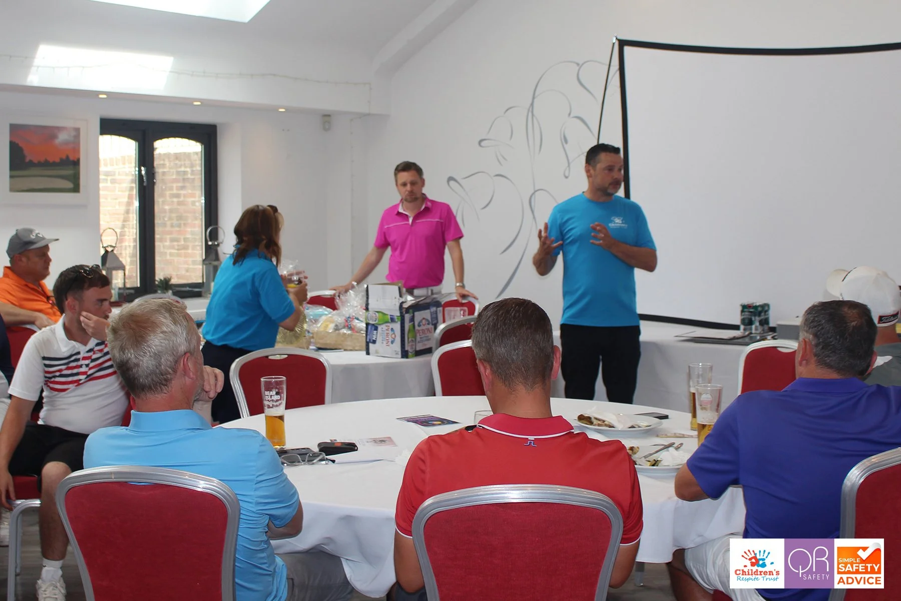 A man in a blue T-shirt is speaking to a group of people seated at round tables in a bright room with white walls and windows. Some people are holding drinks, and there are snacks and miscellaneous items on the tables. A woman in a blue shirt and two