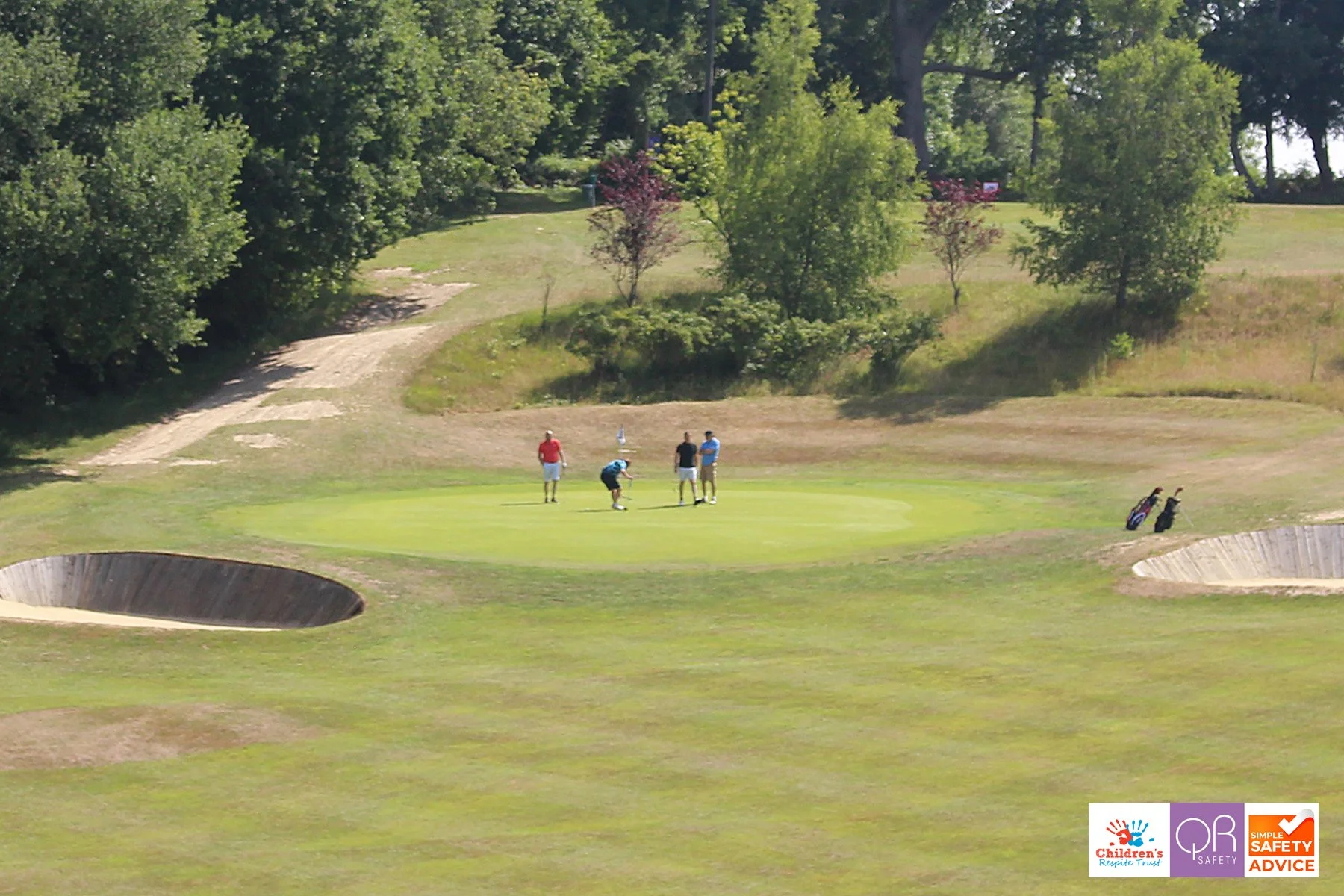Four people on a golf course near a putting green, surrounded by trees and sand bunkers.