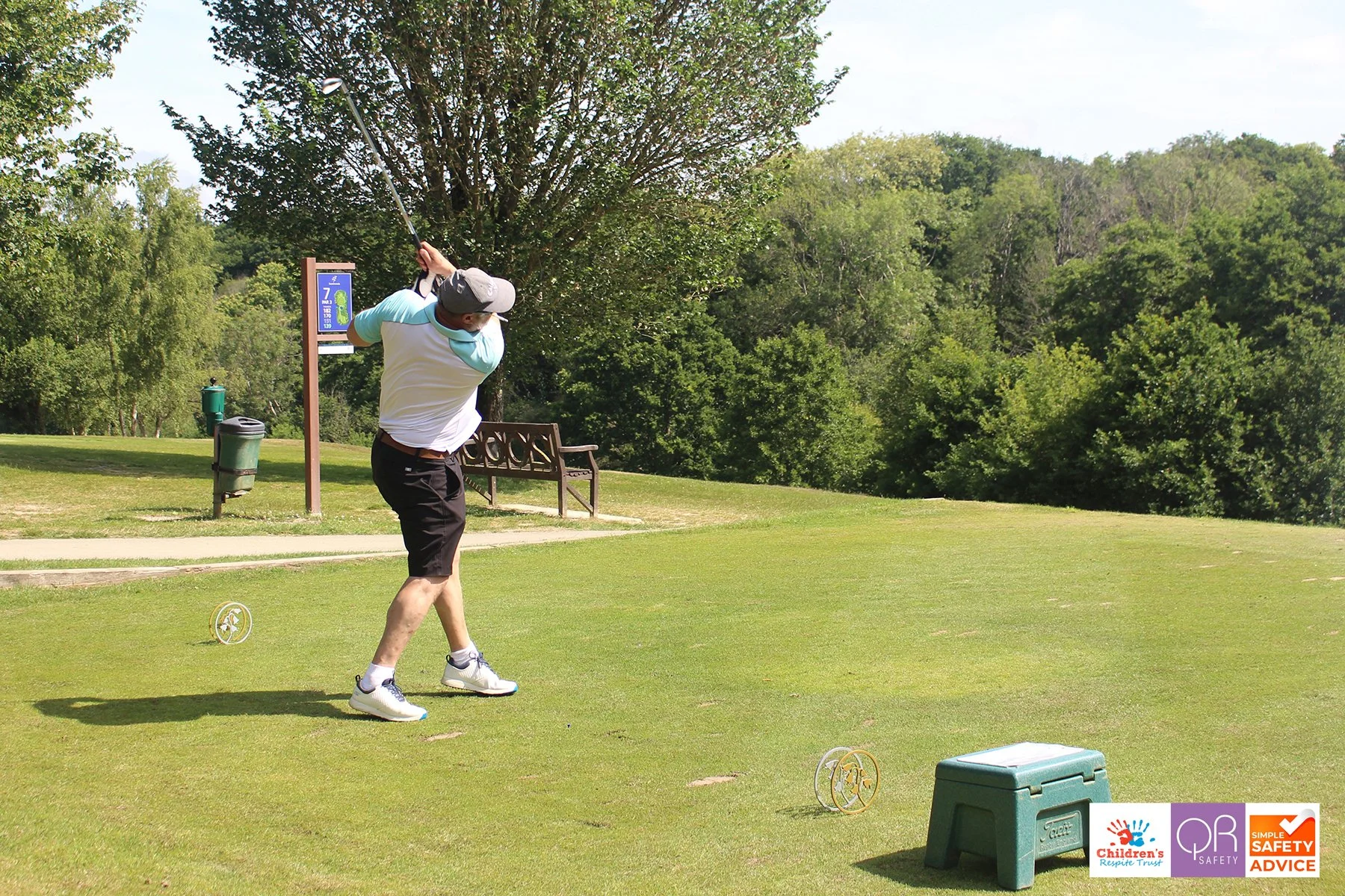 A man practicing golf on a grassy course, mid-swing with a golf club, surrounded by trees and a bench.