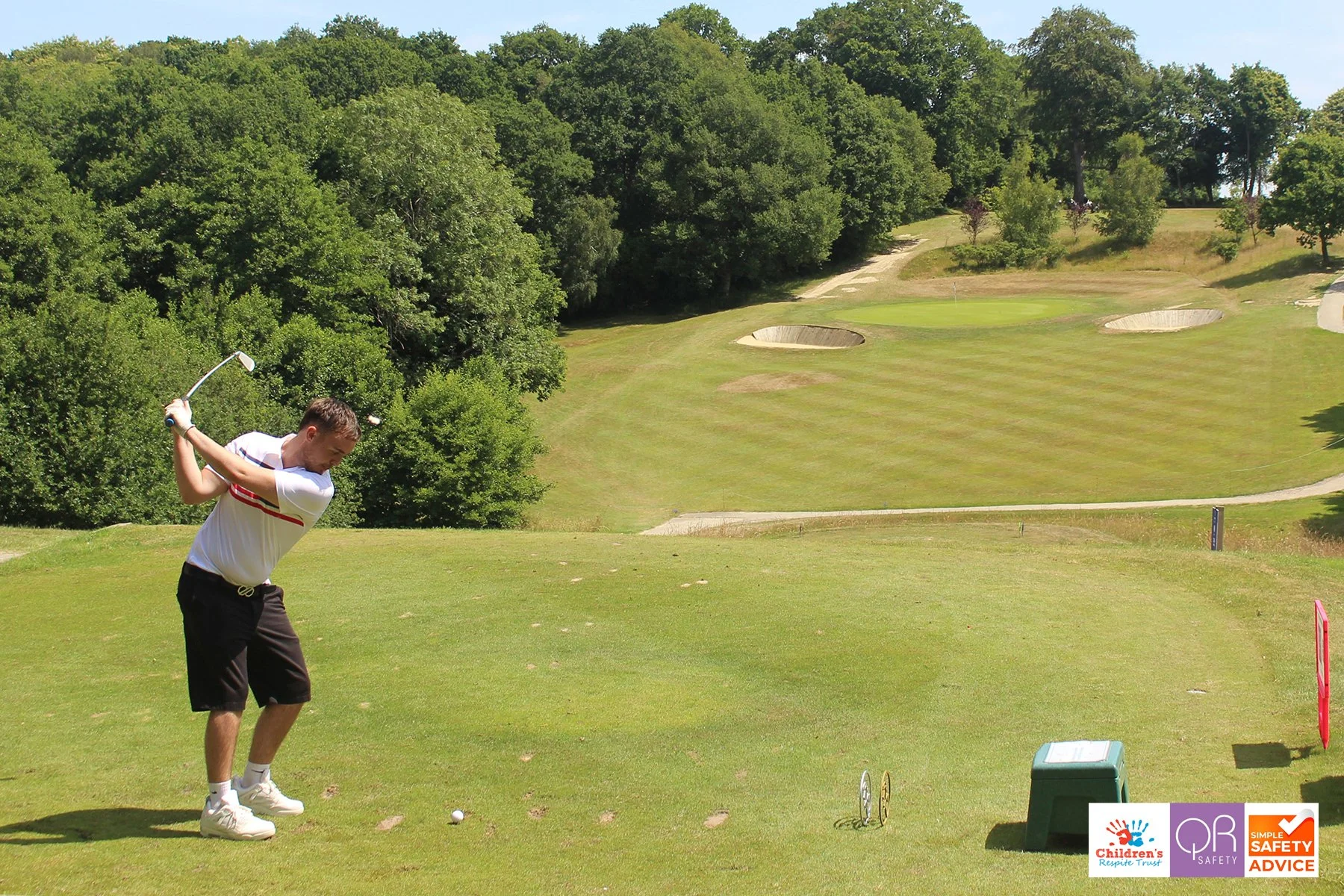 A young man preparing to hit a golf ball on a golf course with trees and multiple bunkers in the background.